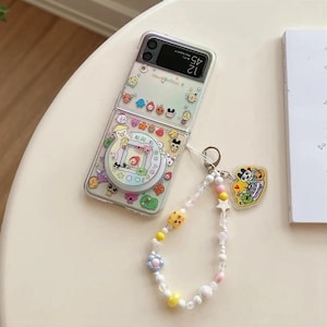 May include: Transparent phone case adorned with cartoon characters and a pop socket with a retro game design. A beaded charm bracelet with star and flower details is attached. The phone is open, showing the screen.