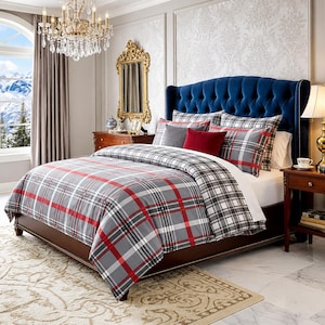 May include: A bed with a blue, buttoned headboard and a plaid duvet cover in grey, red, and white. The bed is made up with matching pillows. A gold mirror and a crystal chandelier are also visible in the bedroom.