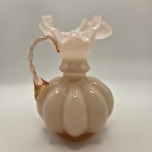 May include: A pale pink glass pitcher with a bulbous, ribbed body and a ruffled rim. The pitcher features a curved handle with a textured, bead-like design and a smooth, translucent finish that catches the light, creating soft highlights.