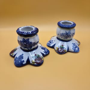 May include: Two white ceramic candle holders with a scalloped base and a cylindrical top. Each piece features hand-painted blue floral patterns and small red and yellow accents around the base and neck, with a dark blue rim at the top opening.