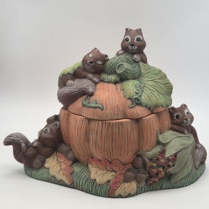 May include: A ceramic lidded jar shaped like a pumpkin, featuring three brown squirrels with painted details. The pumpkin is orange with green leaves, and the base includes sculpted autumn leaves, grass, and a small ear of corn in shades of brown and green.