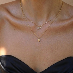 Puede incluir: Dos collares dorados superpuestos en el cuello de una persona. El collar superior tiene un pequeño colgante con la letra "J" y una pequeña gema. El collar inferior tiene un colgante rectangular. La persona lleva una parte superior negra.