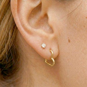 Pode incluir: Close-up de uma orelha com dois brincos. Um é um pequeno e transparente ponto. O outro é um brinco de argola dourado em forma de coração. Pele clara.