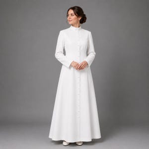 May include: A long, white dress with a high collar and long sleeves. The dress is buttoned down the front and flares out from the waist. The woman is wearing white shoes. The background is a solid gray.