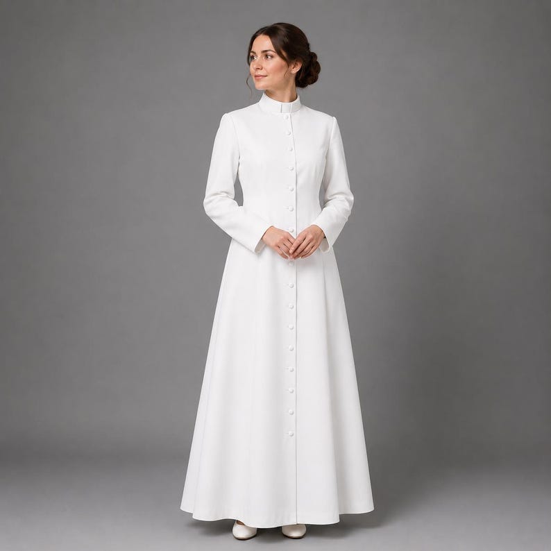 May include: A long, white dress with a high collar and long sleeves. The dress is buttoned down the front and flares out from the waist. The woman is wearing white shoes. The background is a solid gray.