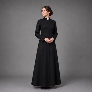 May include: A long, black dress with a high, rounded collar and long sleeves. The dress is buttoned down the front and flares out from the waist. A white collar is visible at the neck. The dress is worn with black shoes.