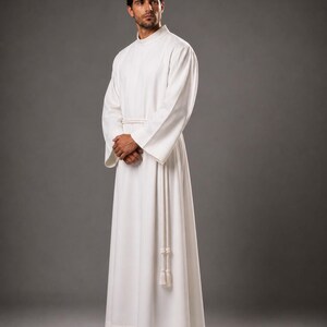 May include: A white, long-sleeved robe with a high collar and a matching rope belt. The robe is floor-length and has wide sleeves. The person is wearing black shoes. The background is a solid gray.