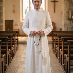 Puede incluir: Un hombre con una túnica clerical blanca está de pie en una iglesia. La túnica tiene cuello alto y botones en la parte delantera. Sostiene un rosario con una cruz. El fondo incluye bancos de madera, una vidriera y un crucifijo.