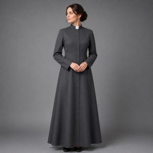 May include: A long, charcoal gray dress with a high, white collar and long sleeves. The dress features a row of buttons down the front and a flared skirt. The woman is wearing black shoes. The dress is a classic, formal style.