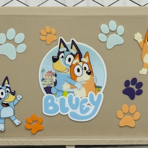 Può includere: Un contenitore beige decorato con i personaggi di Bluey e Bingo, impronte di zampe e la parola "Bluey". I personaggi sono in varie pose, con accenti blu, arancioni e bianchi.