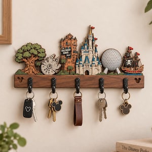 Può includere: Un portachiavi da parete in legno con un design a strati ritagliati di monumenti di parchi a tema, tra cui un albero, un'astronave, l'Hollywood Tower Hotel, un castello, una cupola geodetica e una nave pirata. Cinque ganci neri reggono le chiavi.