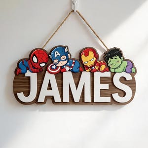 May include: A wooden wall hanging sign featuring the name JAMES in large white letters. Above the name are four cartoon superhero figures: Spider-Man, Captain America, Iron Man, and the Hulk, all in a sleeping pose. The sign hangs from a brown rope.