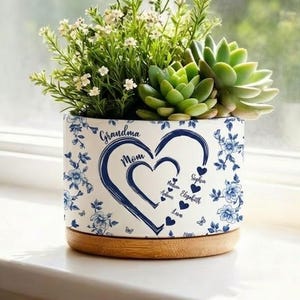 May include: A white ceramic planter with a blue floral design and a wooden base, filled with succulents and small white flowers. The planter features a heart design with the words "Grandma" and "Mom".