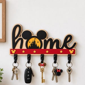 Puede incluir: Un soporte de pared de madera para llaves con la palabra home en letra cursiva negra, con una silueta de castillo dentro de la letra o. La base es roja con estrellas amarillas e iconos de Mickey Mouse, e incluye cinco ganchos negros con llaves.
