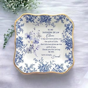 Peut inclure: Un plat carré en céramique blanche avec un bord doré et des motifs floraux bleus. Le plat contient un message à une belle-fille, avec le nom "Olivia" et la signature "Sarah".