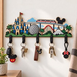 Peut inclure: Un porte-clés mural en bois illustrant une ligne d'horizon de parc à thème avec un château, un monorail, une sphère géodésique et des montagnes russes. Il comporte cinq crochets en métal noir, chacun portant un trousseau de clés avec divers breloques.