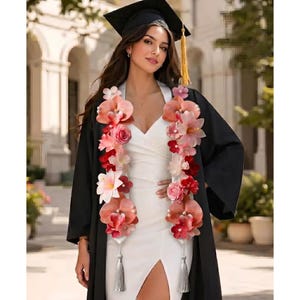 May include: A woman wearing a black graduation gown and cap, adorned with a floral lei. The lei features pink, red, and white flowers. She is wearing a white dress with a high slit. A gold tassel hangs from the cap.