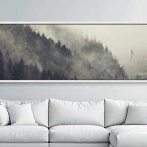 May include: A modern living room featuring a large white sectional sofa, a framed landscape art piece, and a potted plant. The artwork depicts a misty mountain scene. The room has a minimalist design with a neutral color palette.