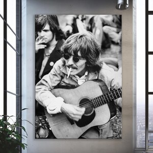 May include: Monochrome photograph of a man playing an acoustic guitar, wearing sunglasses and a light-coloured jacket. Another man is in the background. The image is a vintage style print.