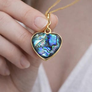 Puede incluir: Collar dorado con colgante en forma de corazón. El colgante presenta una concha de abulón iridiscente, mostrando tonos azules, verdes y morados. La concha está engastada en un marco dorado.