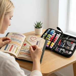 May include: A person is working on a cross-stitch project, with a book open to show various stitch patterns. A black zippered case holds spools of colorful thread, scissors, and other sewing tools. A small succulent plant sits nearby on a wooden table.