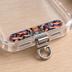 May include: A clear phone case featuring a rectangular graphic insert with a cartoon character surrounded by reaching hands against a dark blue brick background. The case includes a silver metal swivel D-ring attachment at the bottom edge.
