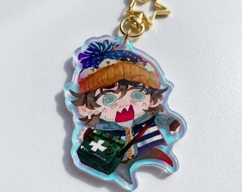 May include: An acrylic keychain featuring a cartoon character with brown hair, wearing a blue and orange beanie, and a blue jacket. The character has a surprised expression and carries a green medical bag with a white cross. It has a gold star clasp.