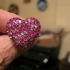 Pode incluir: Um anel em forma de coração com uma superfície coberta por pequenas pedras preciosas redondas rosa e brancas. A banda de metal do anel é visível. As pedras preciosas estão dispostas próximas umas das outras, criando uma aparência texturizada. O anel é uma joia.