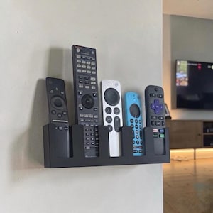May include: A black wall-mounted remote control organizer holding five different remote controls. The remotes are various colors, including black, white, and blue. The organizer is mounted on a white wall.