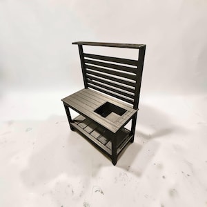 May include: A dark brown wooden mud kitchen toy featuring a slatted back panel, a flat countertop surface with a square cutout basin, and a lower storage shelf. The structure is built with horizontal wooden slats and four sturdy legs.