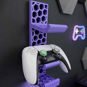May include: A purple wall-mounted gaming controller holder with a honeycomb design, holding a white and black controller. The holder has the text "HEXA-STAND" on it. The background is a dark gray wall with geometric shapes.