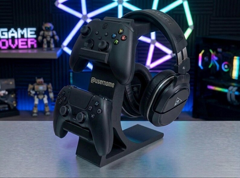 May include: Black gaming headset and controller stand. The stand holds two game controllers and a headset. The stand has the username "@username" printed on it. The background has a "GAME OVER" sign and other gaming accessories.