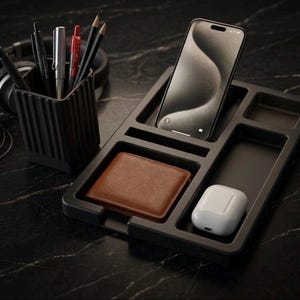 May include: A black desk organizer with compartments for a smartphone, wallet, and wireless earbuds. A black pen holder with pens and pencils is next to the organizer. Headphones are in the background.