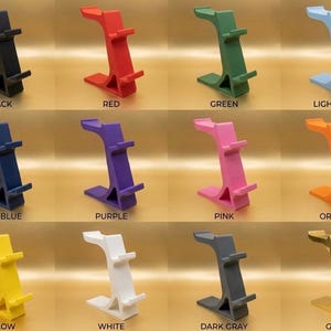 May include: A collection of colorful phone stands in various shades, including black, red, green, light blue, dark blue, purple, pink, orange, yellow, white, dark gray, and gold. Each stand has a unique, curved design.