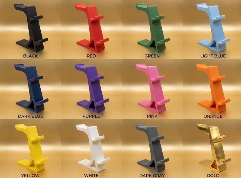 May include: A collection of colorful phone stands in various shades, including black, red, green, light blue, dark blue, purple, pink, orange, yellow, white, dark gray, and gold. Each stand has a unique, curved design.