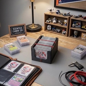 May include: A black plastic trading card storage box with red dividers, holding a stack of cards with a 151 logo. The box is surrounded by several stacks of graded trading cards in plastic cases, a brown leather binder, and a digital caliper.