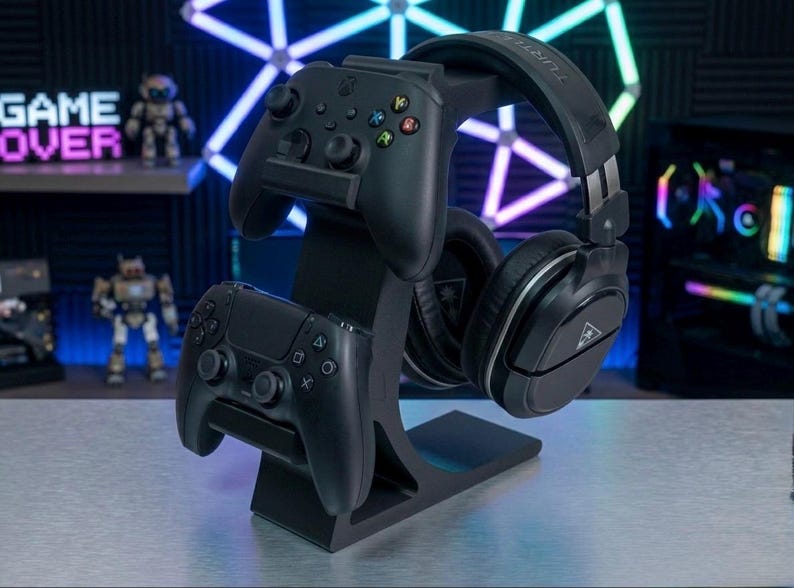 May include: Black gaming headset and two black game controllers on a black stand. The stand is designed to hold the headset and controllers, with the controllers stacked on top of each other. The background includes a shelf with the words "GAME OVER".