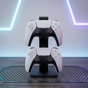 May include: A black vertical stand holding two white and black wireless game controllers stacked one above the other. The stand features a square base and a central pillar designed to securely support the gaming peripherals in a vertical orientation.