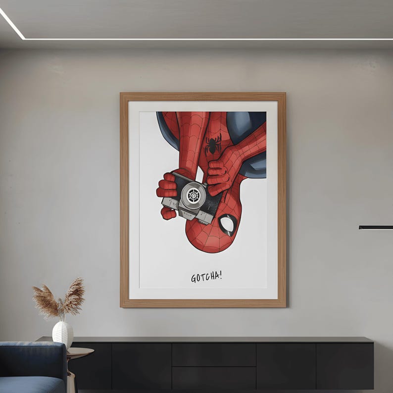 May include: A framed print featuring Spider-Man holding a camera, with the text "GOTCHA!" below. The artwork depicts Spider-Man in his red and blue suit, with a detailed illustration style. The frame is a light brown color.