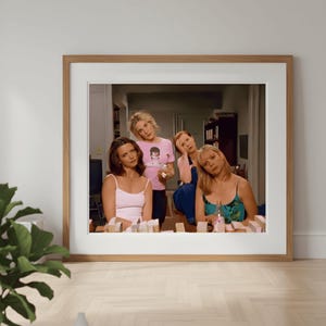 Puede incluir: Una impresión enmarcada que muestra a cinco mujeres en una habitación. Una mujer lleva una camiseta rosa con un gráfico. Las mujeres están rodeadas de pequeños objetos rectangulares. El marco es marrón claro.