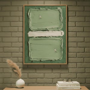 May include: Abstract art piece in a wooden frame. The artwork features a textured, light green background with two rectangular shapes and a white horizontal bar. Tiny figures are placed on the green shapes. The artwork is displayed on a brick wall.