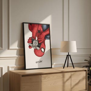 May include: Framed art print featuring Spider-Man holding a camera, with the text "GOTCHA!". The artwork depicts the superhero in his classic red and blue suit, with a black frame. The print is displayed on a wooden cabinet.