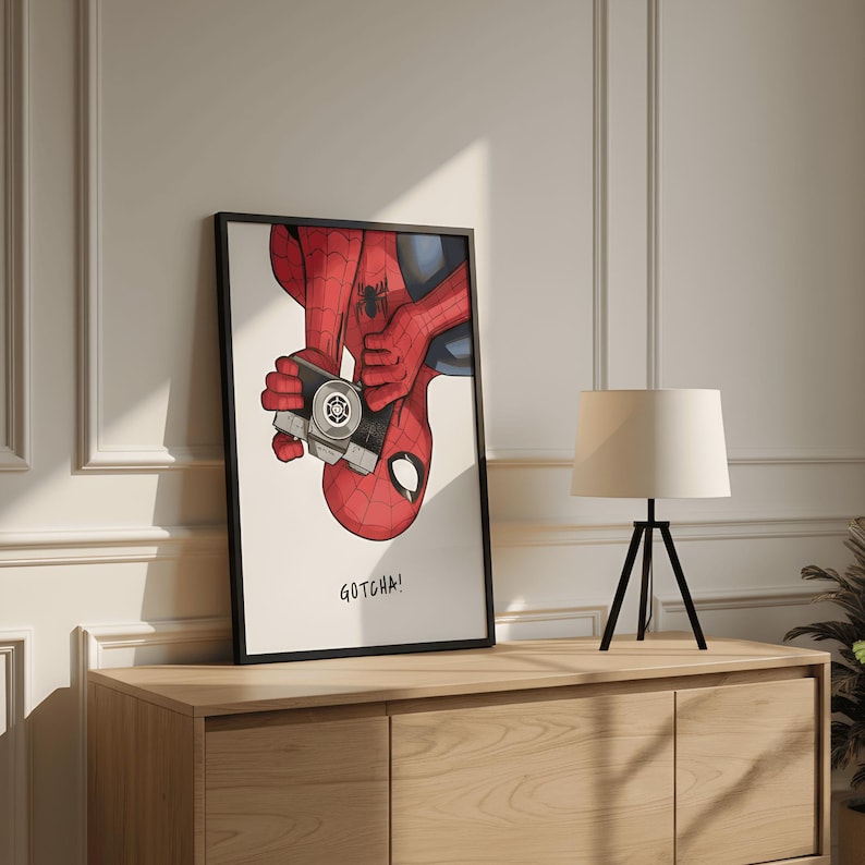 May include: Framed art print featuring Spider-Man holding a camera, with the text "GOTCHA!". The artwork depicts the superhero in his classic red and blue suit, with a black frame. The print is displayed on a wooden cabinet.