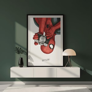 May include: Framed art print featuring Spider-Man holding a camera, with the text "GOTCHA!". The artwork depicts the superhero in his classic red and black suit, with a white background and a black frame. The print is displayed on a white shelf.