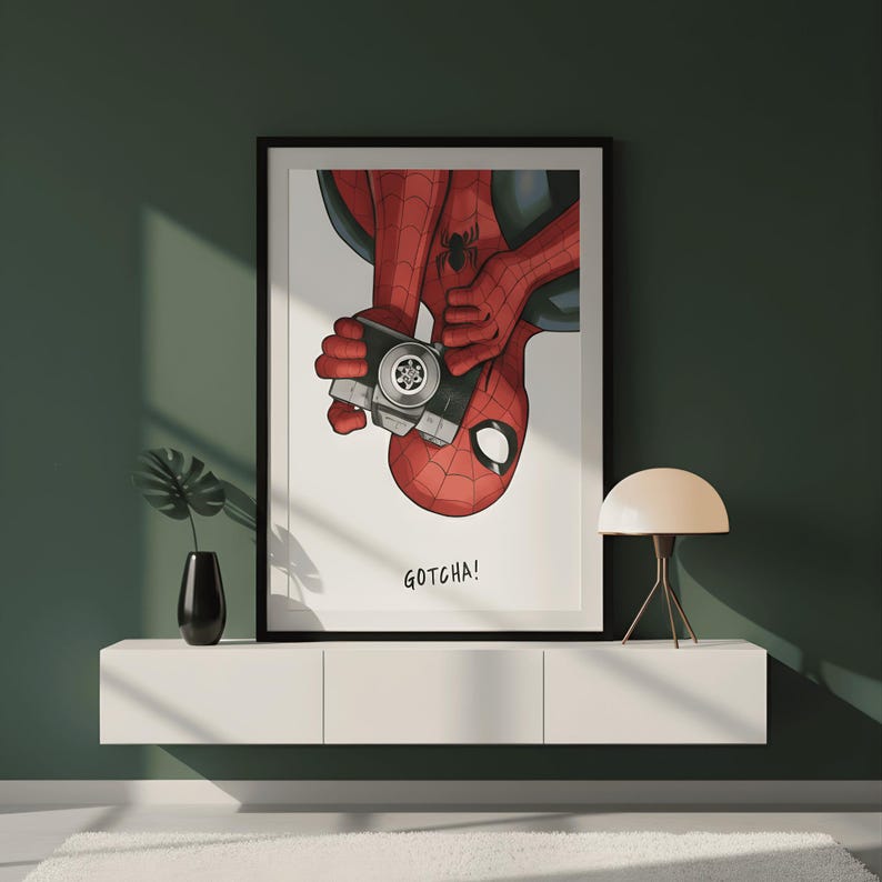May include: Framed art print featuring Spider-Man holding a camera, with the text "GOTCHA!". The artwork depicts the superhero in his classic red and black suit, with a white background and a black frame. The print is displayed on a white shelf.