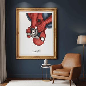 May include: A framed art print featuring Spider-Man holding a vintage camera. The illustration shows Spider-Man upside down, with his red and black suit and the text "GOTCHA!". The frame is gold.