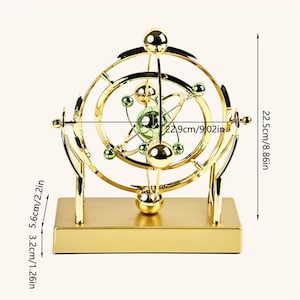 May include: A gold-colored, kinetic art sculpture with a base and a series of interconnected rings and spheres. The sculpture is 9.02 inches wide and 8.86 inches tall. It features green accents and a central, star-like design.