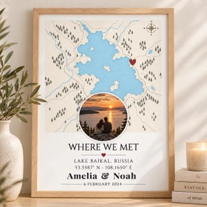 Op de afbeelding: Een ingelijste kaartprint met een lichtblauwe meerillustratie met een rood hartje, een ronde zonsondergangfoto van twee personen en de tekst WHERE WE MET, LAKE BAIKAL, RUSSIA, 53.5587 N 108.1650 E, Amelia & Noah, 6 FEBRUARY 2024.