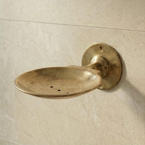 Pode incluir: Um saboneteira de latão de estilo vintage, montada na parede. Possui uma placa traseira circular e um suporte oval para sabão com três orifícios de drenagem. O latão tem uma pátina quente e envelhecida. Para o banheiro.