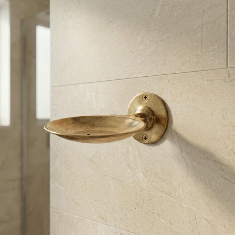 May include: A wall-mounted soap dish made of brass. The dish has a circular base with four screws and a curved, oval-shaped holder. The brass has a slightly aged appearance, and the dish is mounted on a light-colored tiled wall.